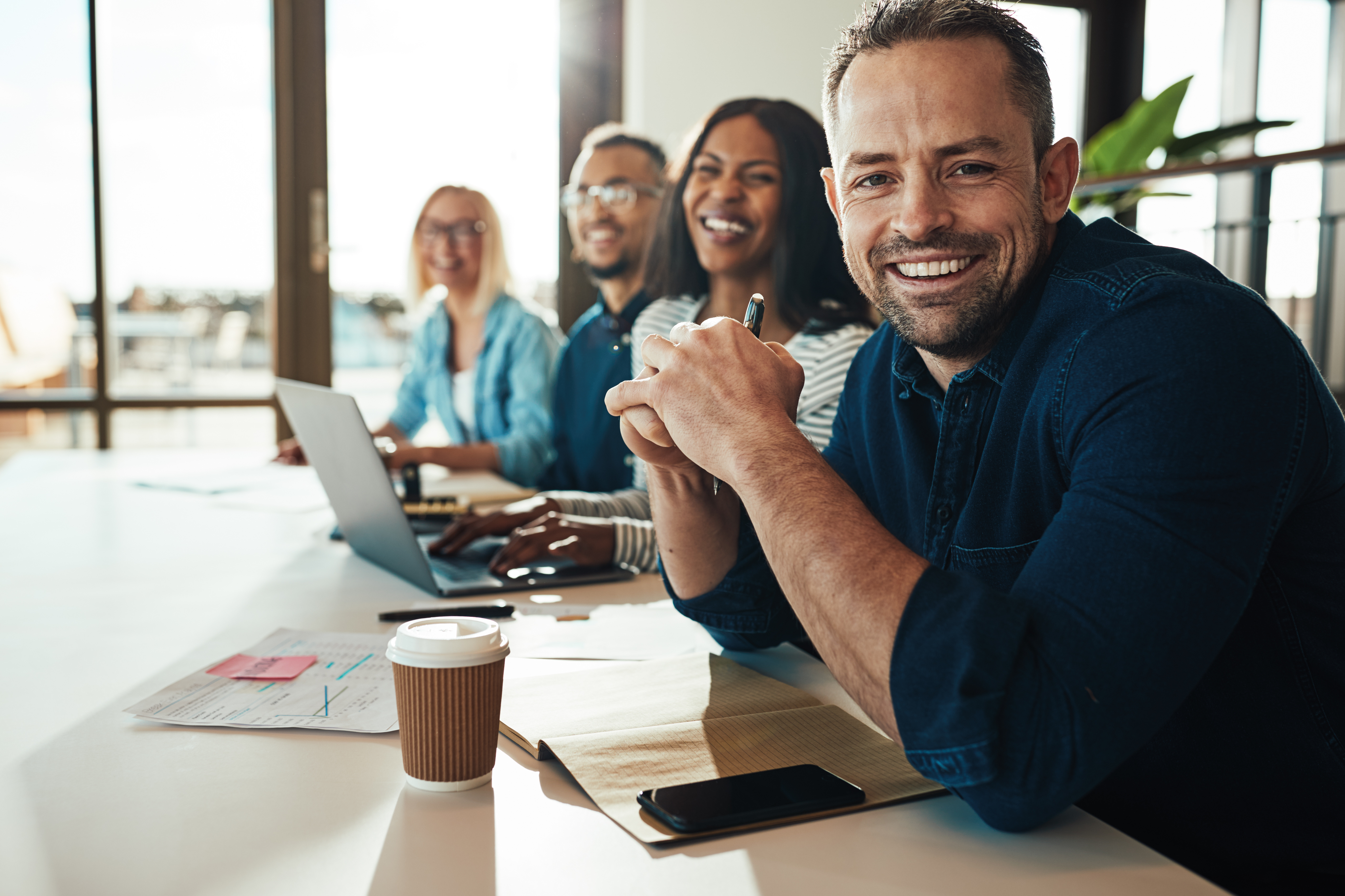 Two women and two men are sitting at a meeting room table with a coffee cup, laptop, phone, and documents and smiling at the camera