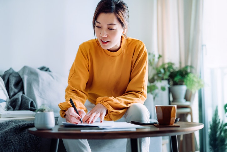 A woman in a yellow sweater is smiling and writing notes at a coffee table with a mug, and a small pot plant