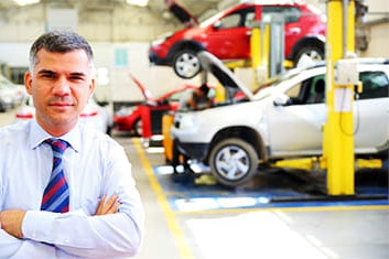 A friendly man in a suit looks at the camera with his arms crossed. Cars are raised up being serviced behind him