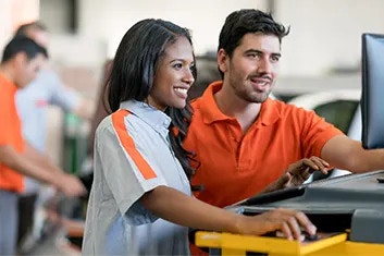 A smiling woman is controlling a computer, and her smiling male colleague is looking at her screen. A vehicle service department is in the background