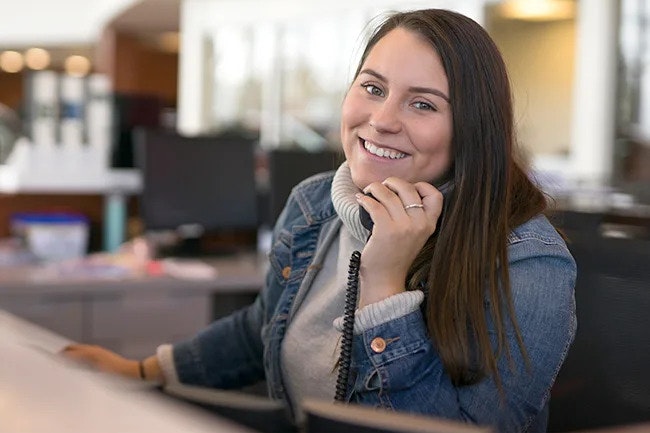 A long-haired woman in a hoodie and denim jacket, sitting in a chair at a reception desk, smiling at the camera