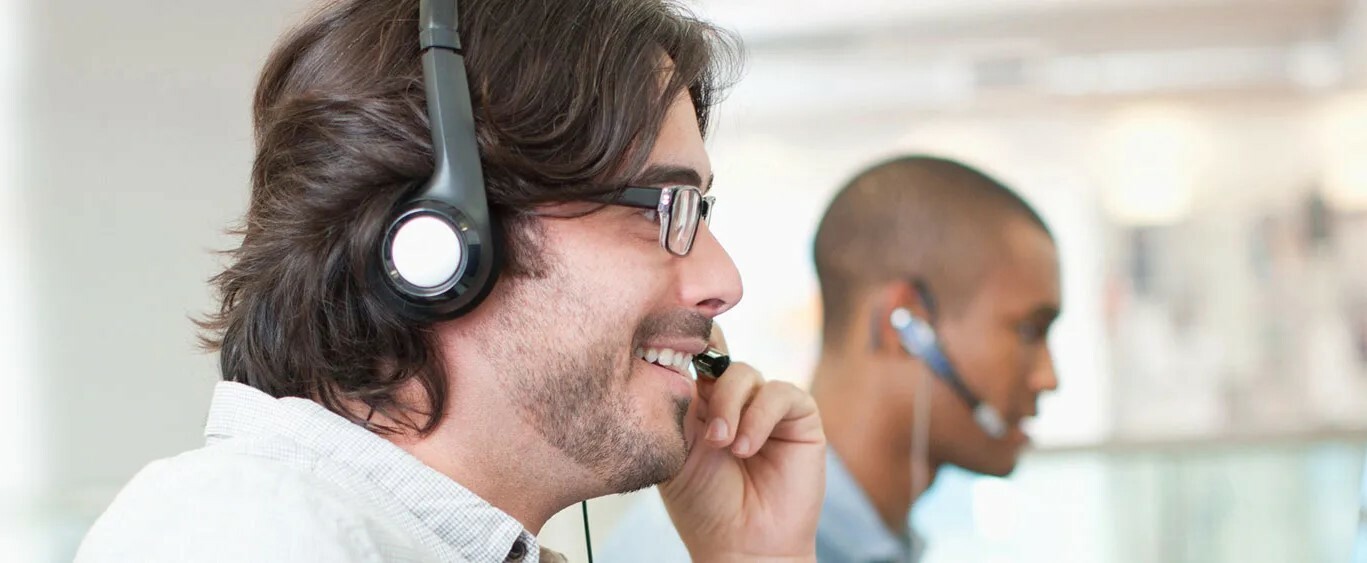 A smiling man talking on a headset in an office. He is facing his computer, and two smiling colleagues are sitting on either side of him