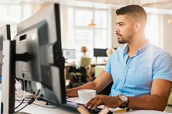 A man in a short-sleeved blue shirt, working in front of his computer at a desk in a bright office