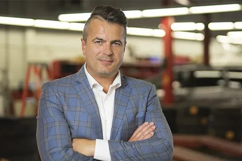 A slightly smiling man is in a plaid suit with his arms crossed, looking at the camera. A dealership is in the background