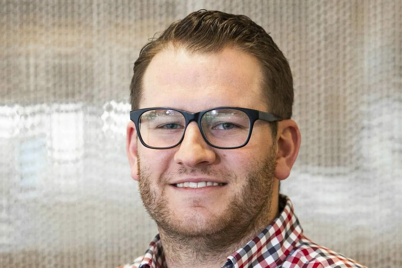 A smiling man with glasses and a beard is wearing a chequered blue, red and white shirt, and looking at the camera