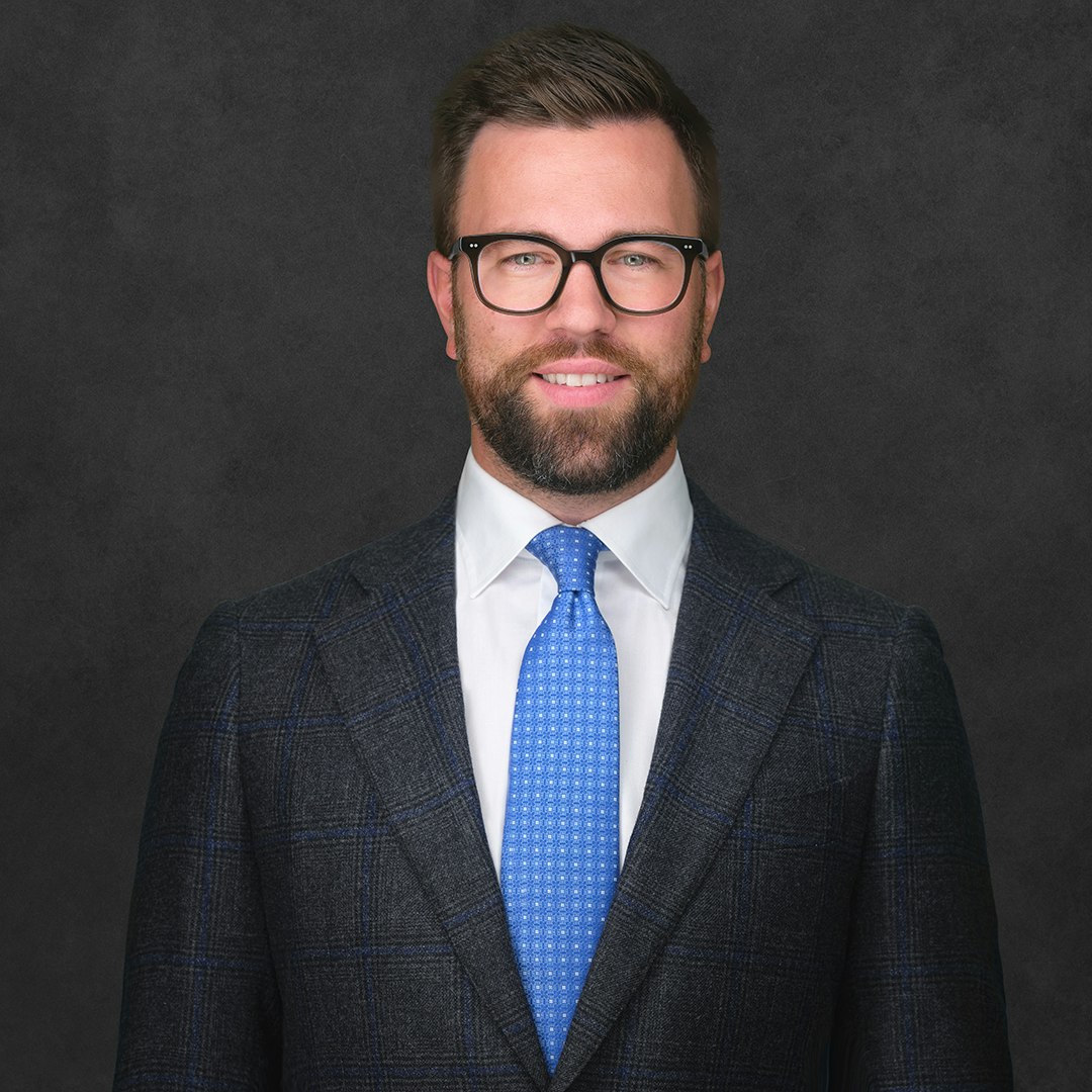 Jared Biggs smiling, wearing glasses, a suit, a white shirt and blue tie