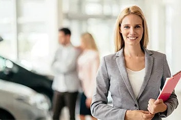 A woman in a suit is holding a red folder and smiling at the camera, two customers are looking at cars behind her