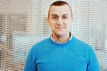 A smiling man in a blue sweater is smiling at the camera. There is a beaded curtain behind him