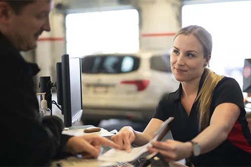 A smiling female worker in a black and red polo shirt shows a document to a smiling man. There are cars in the background