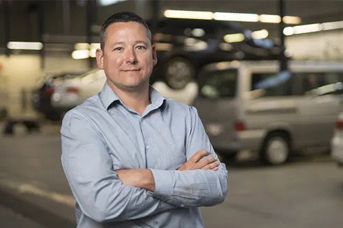 A smiling man in a blue shirt is smiling at the camera, with his arms folded, standing in front of a number of vehicles
