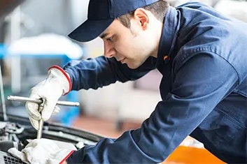 A man in blue overalls and a baseball cap is servicing a vehicle under it's hood